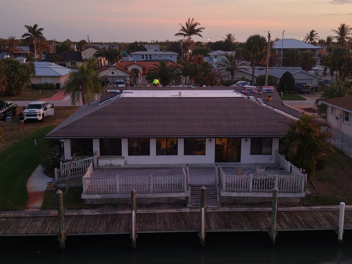 Drone view at dusk of a canal-front home during reroof, crew installing a white TPO flat roof beside shingle hip slopes