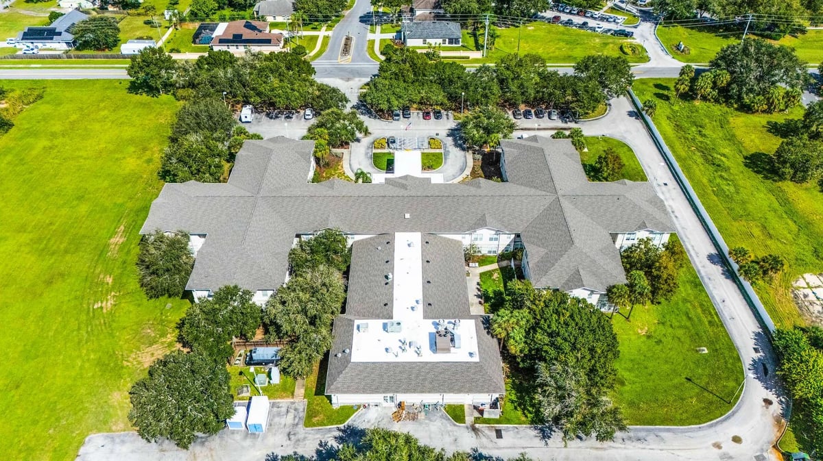 Aerial view of a large modern house with gray roof surrounded by green lawns, landscaping trees, and a driveway in a residential neighborhood