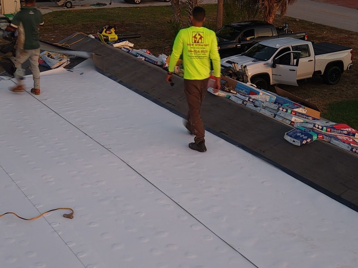 Roofer in a hi-vis company shirt walking on new white TPO flat roof next to a shingle slope with bundles staged