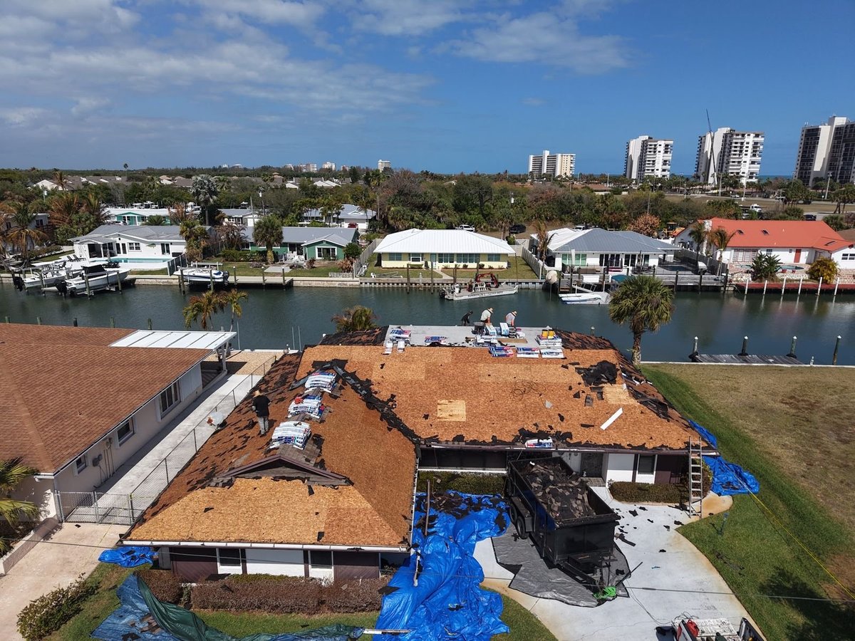 Aerial view of a shingle roof tear-off on a canal-front home, bare decking, shingle bundles staged and tarps protecting the yard