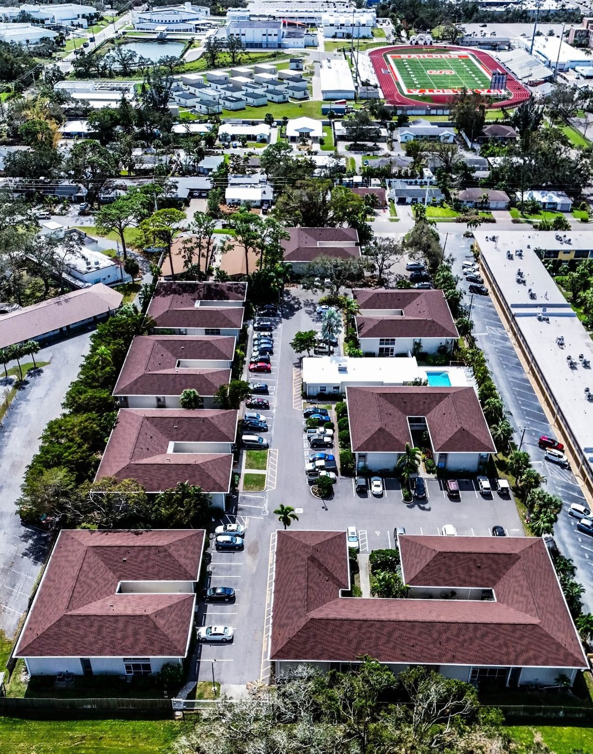 Overhead drone view of red-brown hip shingle roofs lining the parking area of a multifamily condo complex