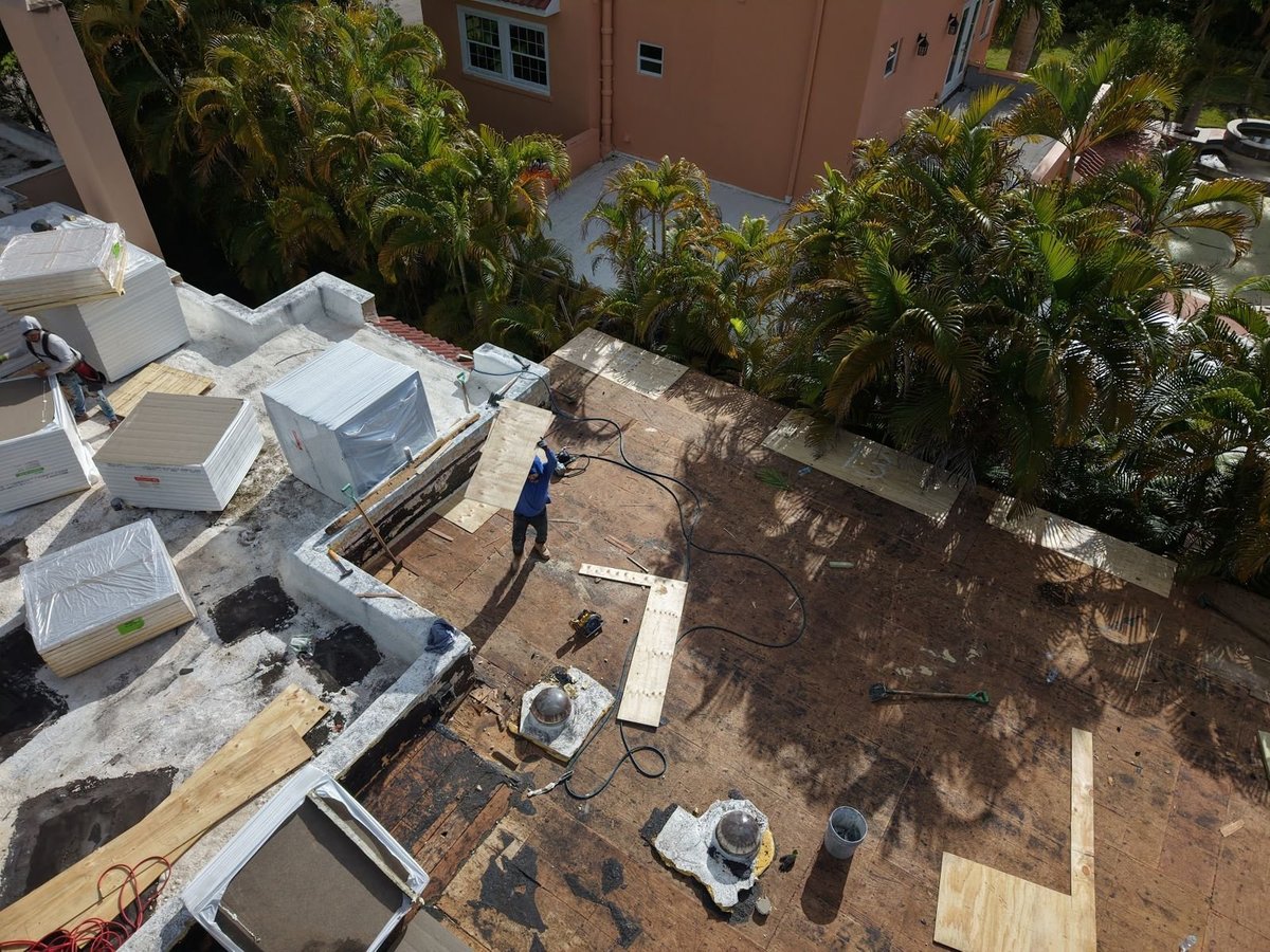 Roofer replacing damaged wood decking with new plywood on a stripped flat roof before insulation is installed